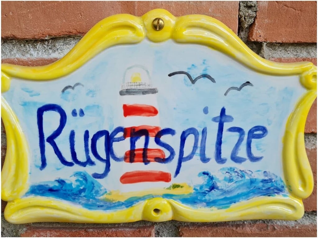 A colorful ceramic sign features the word 'RügenSpitze' painted in vibrant blue letters. A stylized red and white lighthouse is depicted alongside waves and seagulls, set against a light blue background. The sign is framed in a bright yellow border.