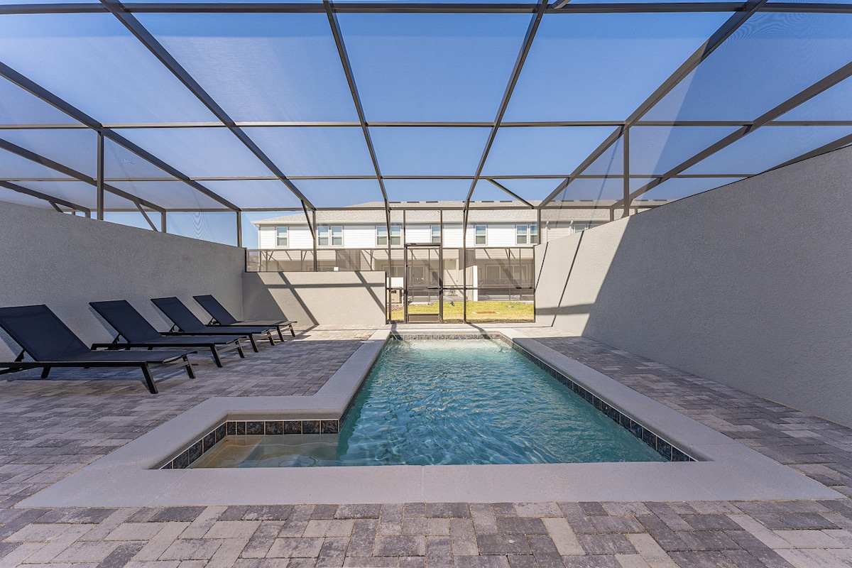 A private pool area is enclosed by a screened lanai, showcasing a clear swimming pool surrounded by paving stones. Several lounge chairs are positioned around the pool, offering a comfortable spot to relax. Natural light streams in, creating a bright and inviting atmosphere.