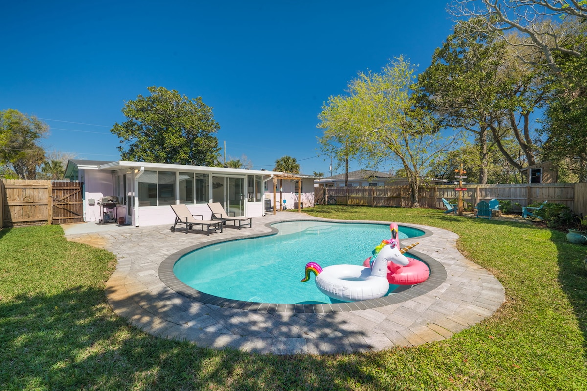 A sparkling saltwater pool is surrounded by a spacious yard, featuring charming lounge chairs on the patio. Colorful floaters in the shape of swans are positioned in the water, offering a playful touch against the vibrant blue sky and well-maintained greenery.