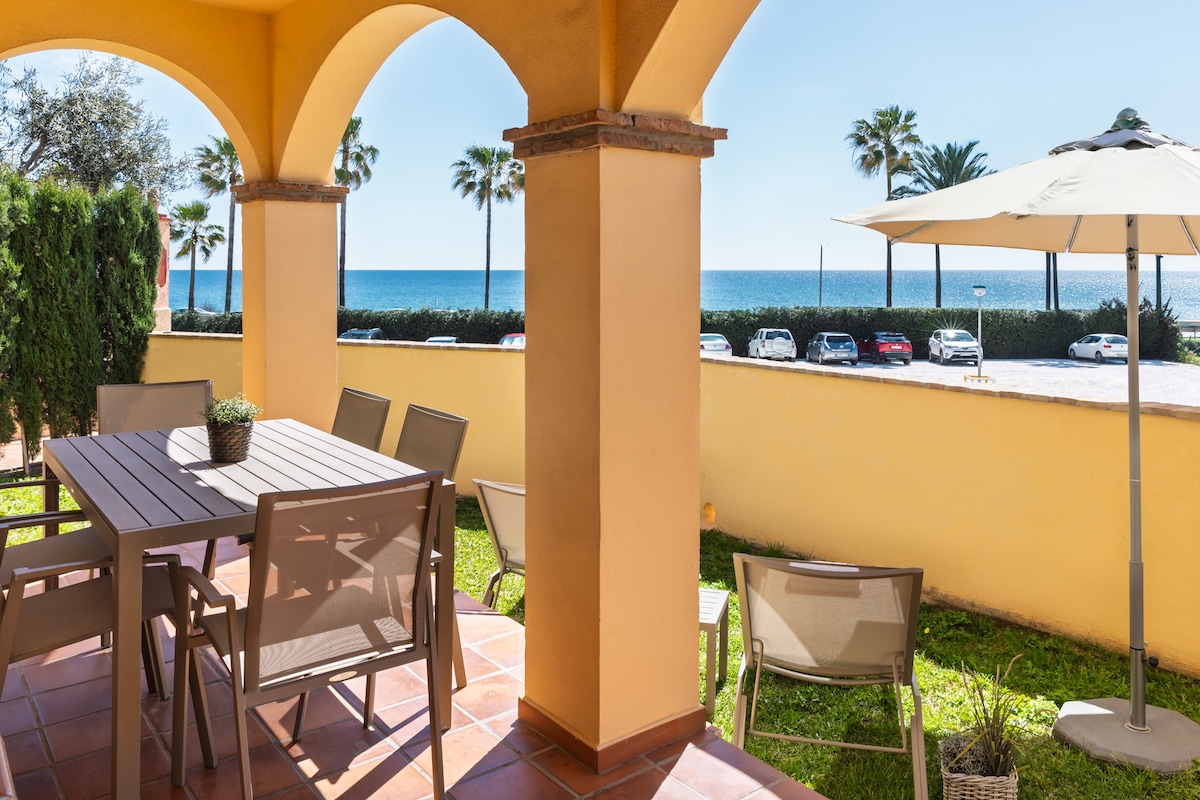 A terrace features a dining table surrounded by several chairs, with a large umbrella providing shade. Lush greenery is present, and views of the sea and palm trees are visible in the distance, enhancing the tranquil outdoor setting.