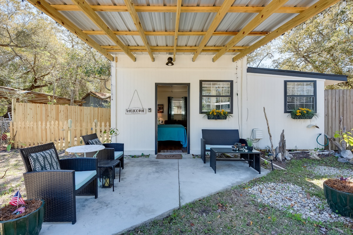 An outdoor seating area is set under a covered porch, featuring a table with four chairs and two lounge sofas. Potted plants add a touch of greenery, while the entrance to the studio is visible in the background, hinting at a cozy interior.