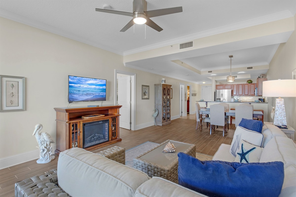 An open living area features a comfortable seating arrangement with neutral-toned couches and accent pillows. A wooden television stand holds a flat-screen TV, while a decorative sculpture adds interest to the space. Natural light enters through large windows, enhancing the inviting ambiance.