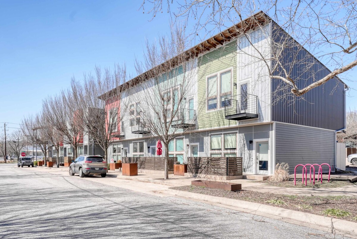 A modern two-story building features a mix of colorful exteriors, including green and red accents. Large windows are complemented by minimalist balconies, and planter boxes are positioned along the walkway. Leafless trees line the street, contributing to a clean and urban atmosphere.