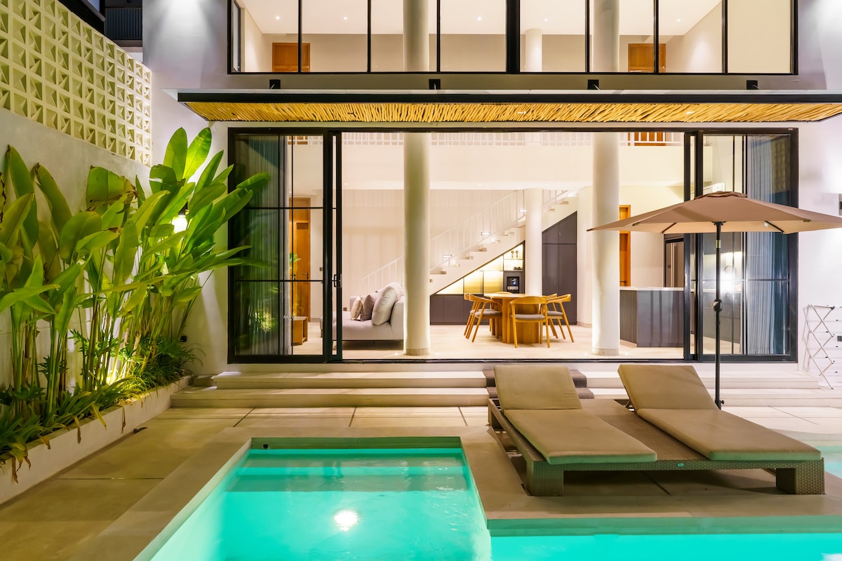 The image showcases a private pool area with two sun loungers positioned beside the water. Large glass doors open to an enclosed living space, revealing a dining area illuminated by warm lights. Lush greenery frames the pool, enhancing the tropical ambiance.