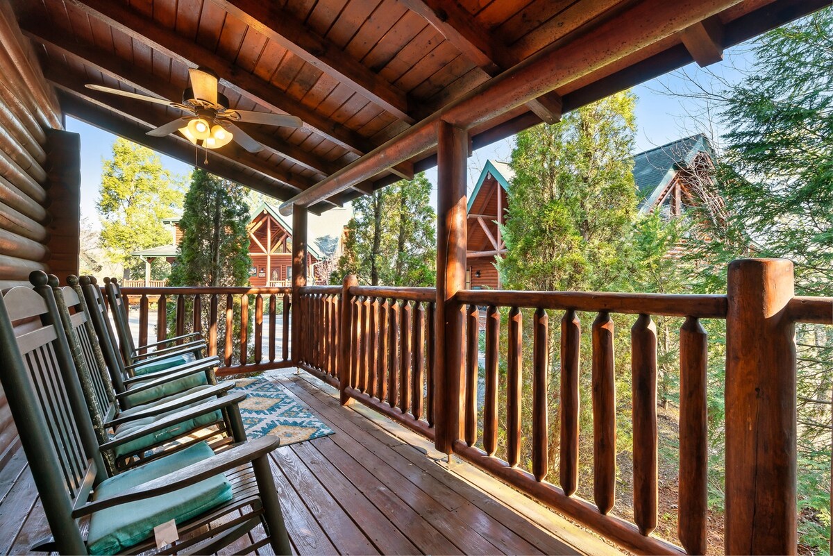 A covered porch with wooden rocking chairs arranged along the railing overlooks a peaceful wooded area. The porch features a wooden ceiling and a rustic feel, with sunlight filtering through the trees nearby. A patterned rug adds a decorative touch to the floor.
