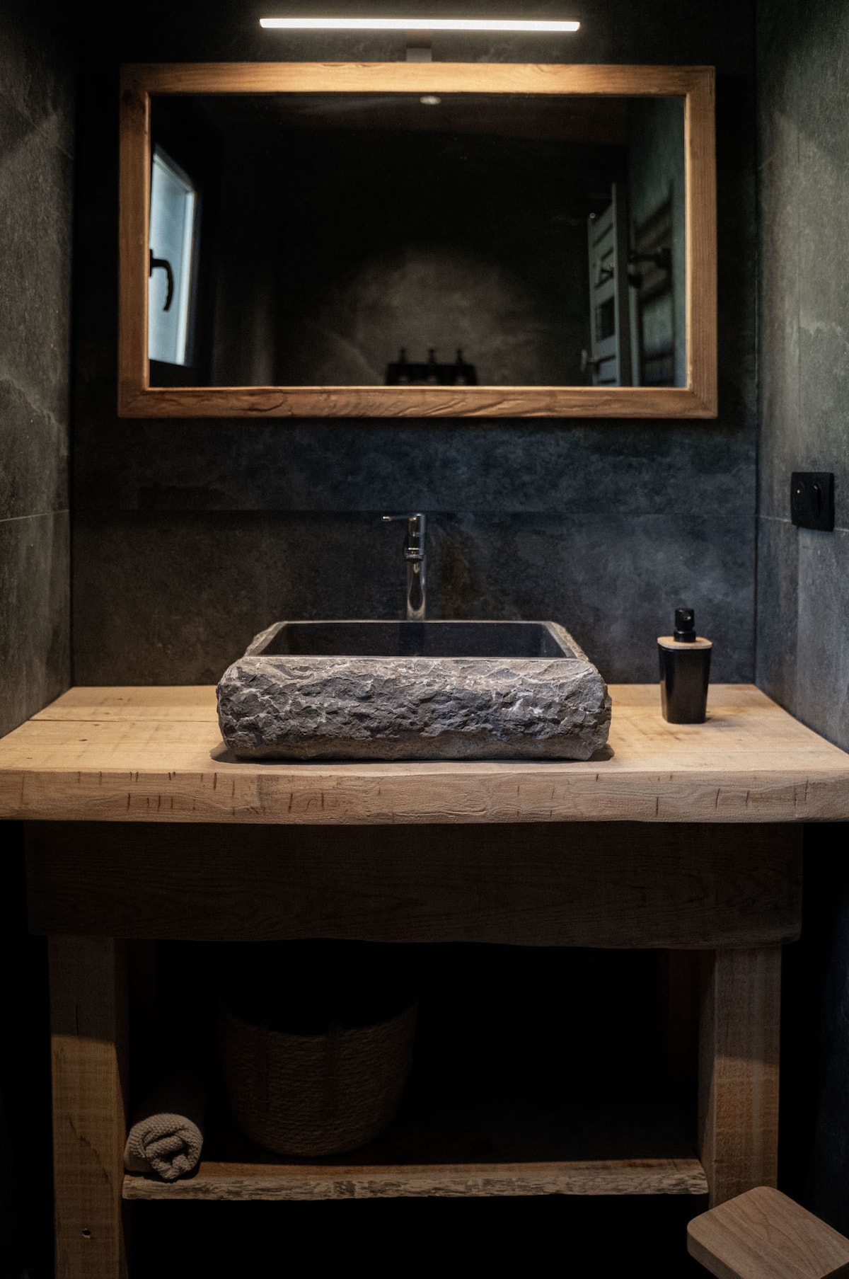 A neutral-toned bathroom features a wooden counter with a stone basin that enhances the natural aesthetic. A large mirror is framed in wood, reflecting the subtle lighting. Below, a wooden shelf is seen, providing space for toiletries and a woven basket.
