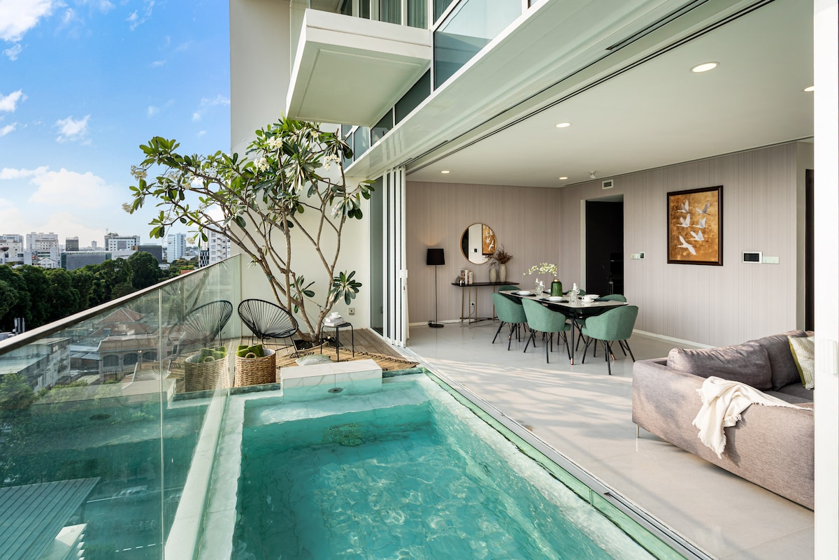 A private balcony is shown with a small pool, surrounded by green plants. The open space flows into a dining area equipped with stylish furnishings, while large glass doors provide unobstructed views of the city skyline and allow natural light to brighten the interior.