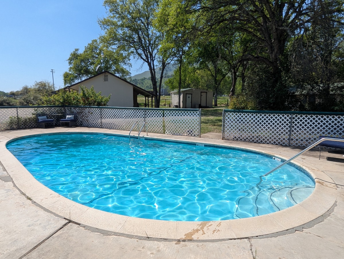 An inviting outdoor swimming pool is surrounded by a spacious deck and a white, lattice-style fence. Lush trees provide shade nearby. Two lounge chairs are positioned beside the pool, offering a relaxing spot for sunbathing. The clear water reflects the sunlight, creating a serene atmosphere.