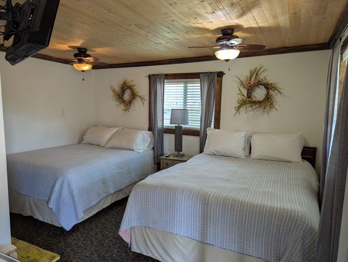 A cozy room is furnished with two spacious beds dressed in light-colored linens. Soft lighting is provided by bedside lamps, and decorative wall accents add character. A window with light-filtering curtains allows natural light to brighten the space.