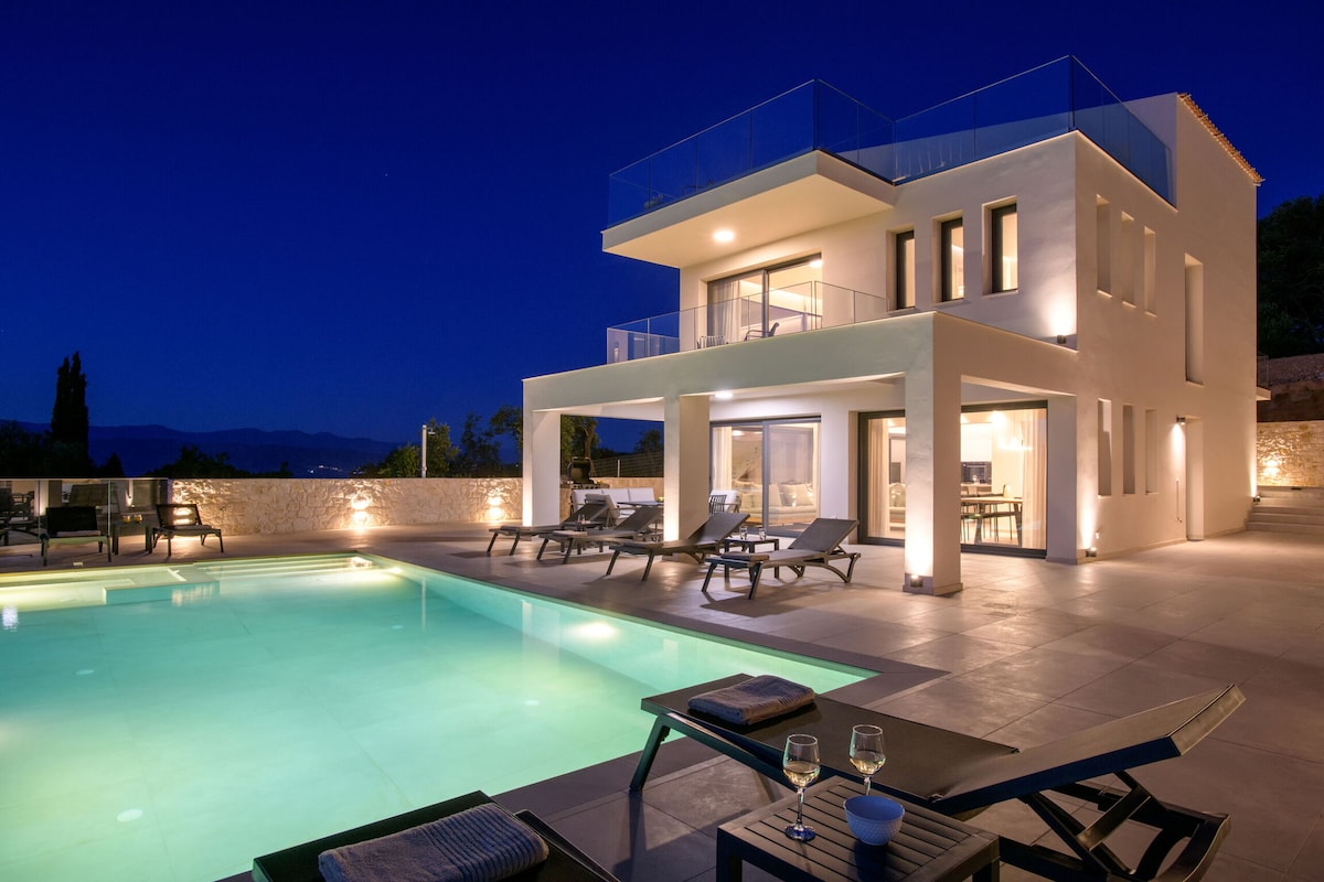 A modern villa exterior presents a private pool illuminated by subtle lighting. The structure features expansive glass doors and multiple terraces. Comfortable lounge chairs are positioned around the pool area, highlighting relaxed outdoor living. Scenic hills are visible in the background, enhancing the tranquil evening setting.