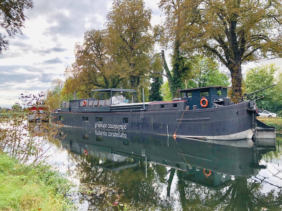 A 1950s barge is anchored along a canal, surrounded by trees. The dark exterior of the barge contrasts with the reflective water. Adjacent foliage creates a serene setting, while the sky above is partly cloudy, adding depth to the tranquil environment.