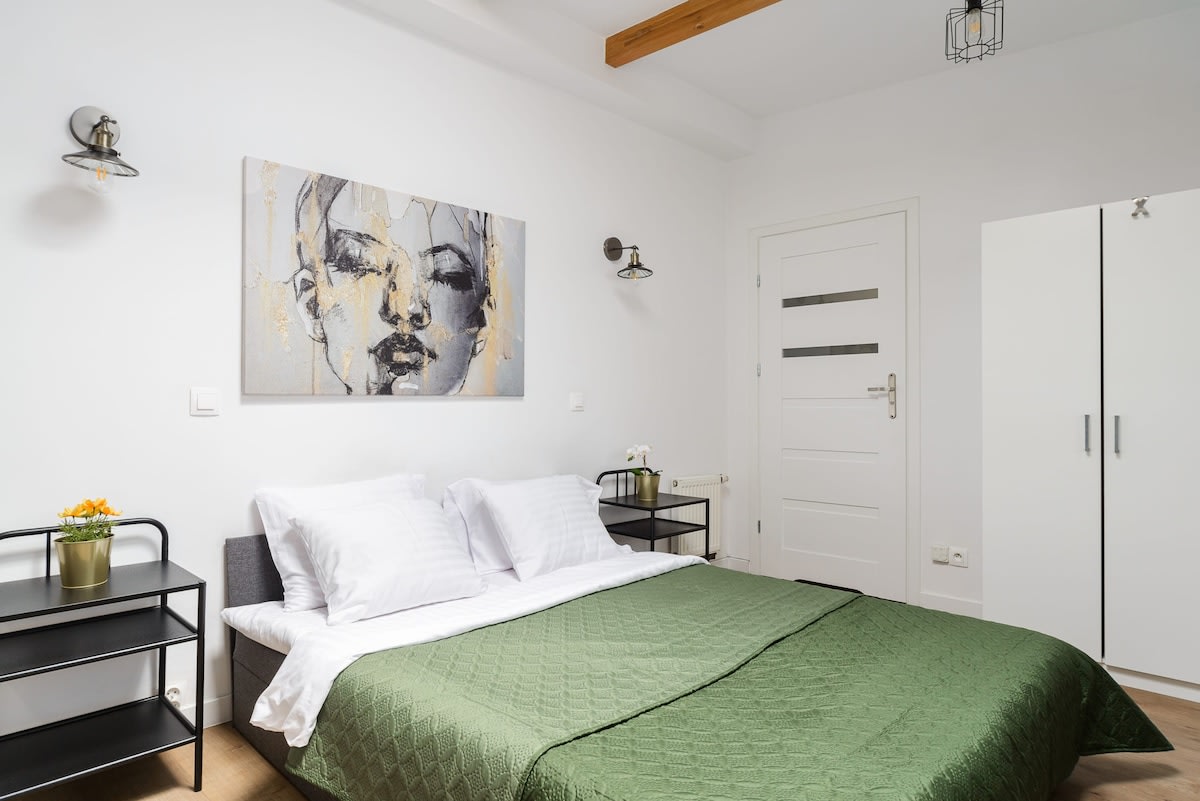 A serene bedroom showcases a spacious bed covered with a green quilt, flanked by black nightstands. Wall sconces provide soft lighting, while modern artwork decorates the wall above. A white wardrobe stands nearby, complementing the light color scheme of the room.