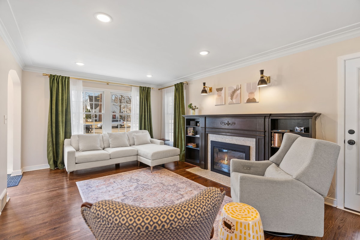 The living room features a neutral color palette with large windows allowing natural light to fill the space. A comfortable sectional sofa is positioned alongside a fireplace, while a patterned area rug lies on the hardwood floor. Decorative wall art and modern lighting add to the inviting atmosphere.