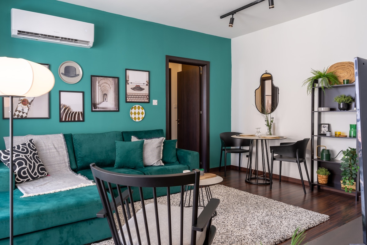 A living room showcases a spacious green sofa adorned with textured cushions, framed artwork on the walls, and a soft area rug. A sleek dining table with black chairs is visible, alongside a modern bookshelf featuring plants and decorative items, creating an inviting atmosphere.