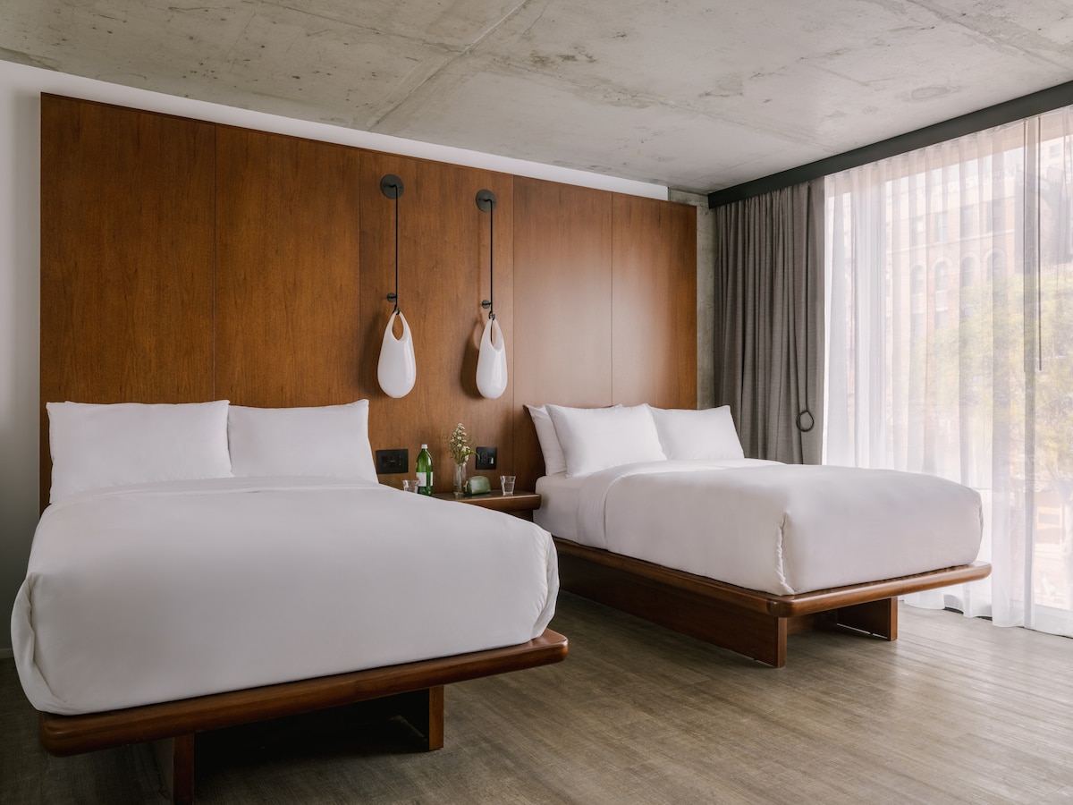 A modern sleeping area features two spacious beds with crisp white bedding, positioned against a stylish wood-paneled wall. Soft natural light filters through floor-to-ceiling curtains, adding warmth to the minimalist decor complemented by sleek bedside lighting and a small side table.
