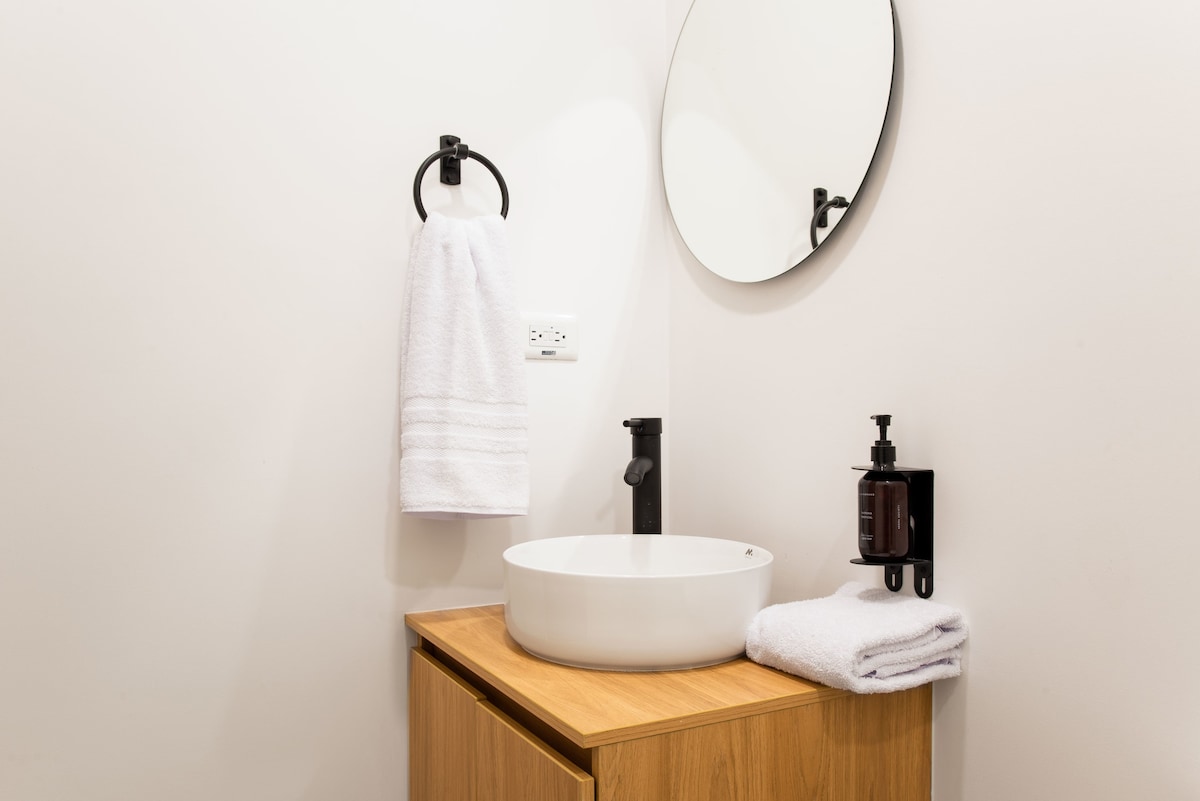 The bathroom features a sleek, circular mirror above a modern sink. A wooden vanity provides storage, complemented by neatly arranged white towels. A wall-mounted soap dispenser adds a touch of convenience, while a minimalist aesthetic enhances the clean design.