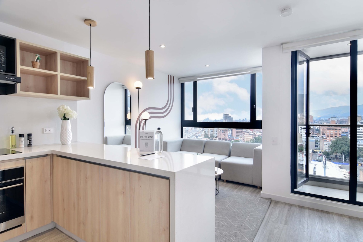 A modern kitchen area is highlighted by light wood cabinetry and a central island. Large windows provide ample natural light and showcase views of the city. A comfortable grey sofa is situated in the adjacent living space, complemented by minimalistic decor.