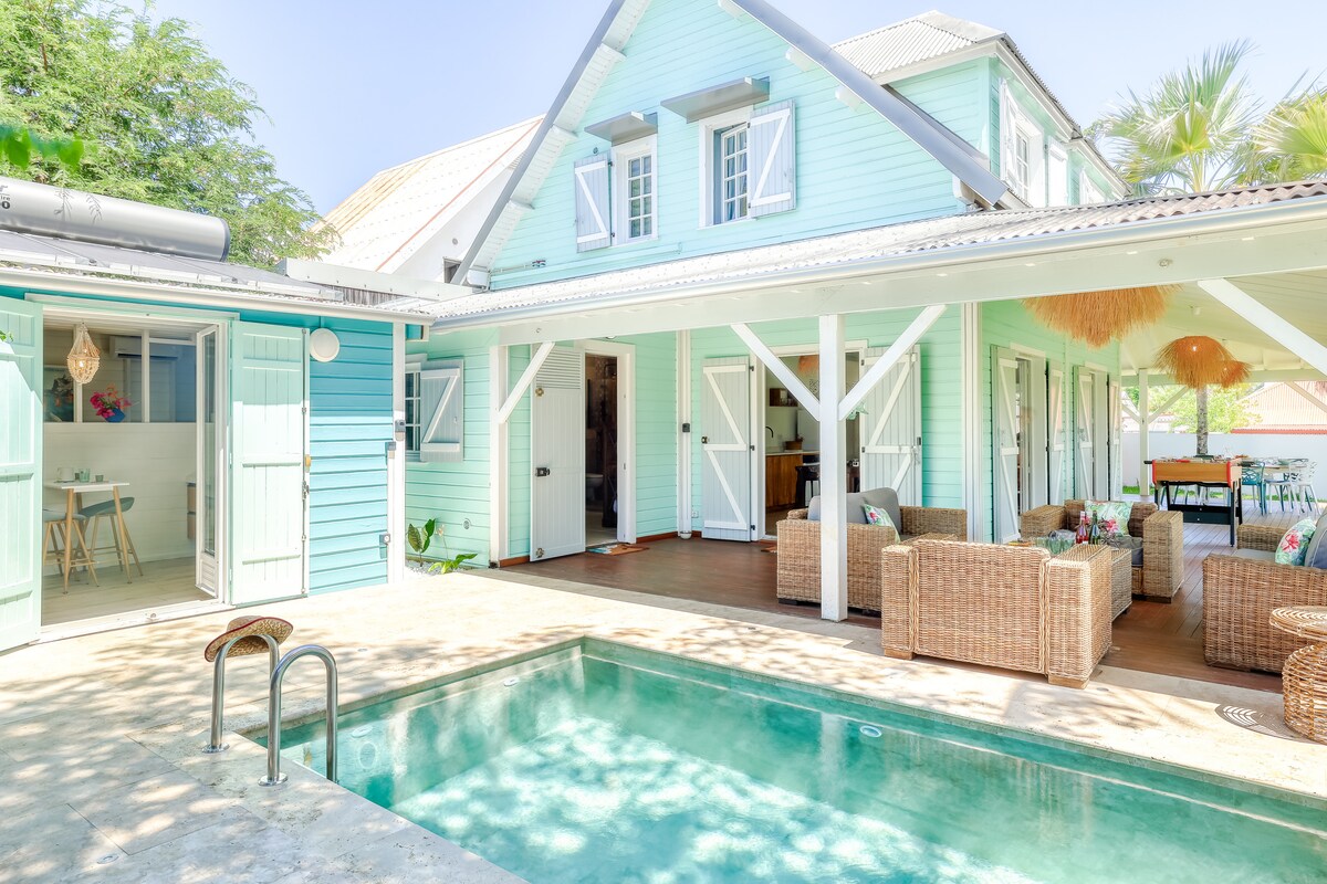 The villa features a private pool with accessible steps, surrounded by a spacious terrace furnished with comfortable seating. The bright exterior showcases a mix of light blue and white finishes, while large doors invite natural light into the open living area connected to an elegant dining space.