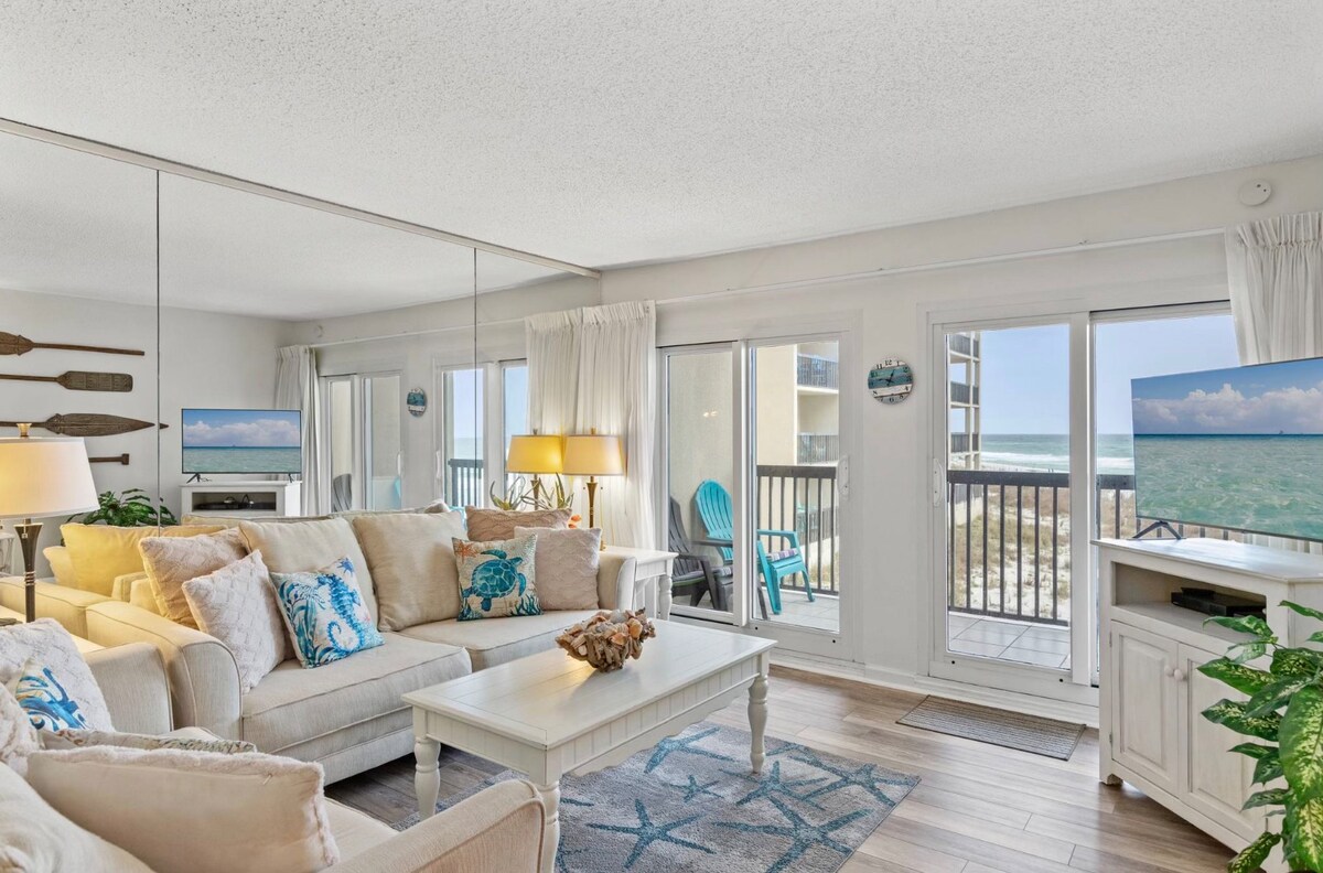 The living area features a comfortable seating arrangement with light-colored sofas adorned with decorative pillows. Large glass doors provide views of the beachfront, allowing natural light to fill the space. A white coffee table is centered in the room, and decorative wall art enhances the coastal theme.