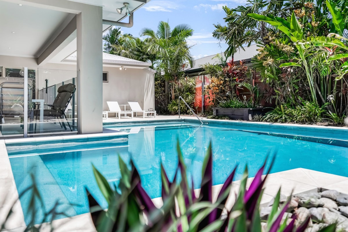 A shimmering swimming pool is framed by lush tropical gardens, showcasing vibrant greenery and colorful plants. Sun loungers are placed nearby, providing a space for relaxation. The surrounding area features an inviting outdoor setting surrounded by palm trees and well-maintained landscaping.