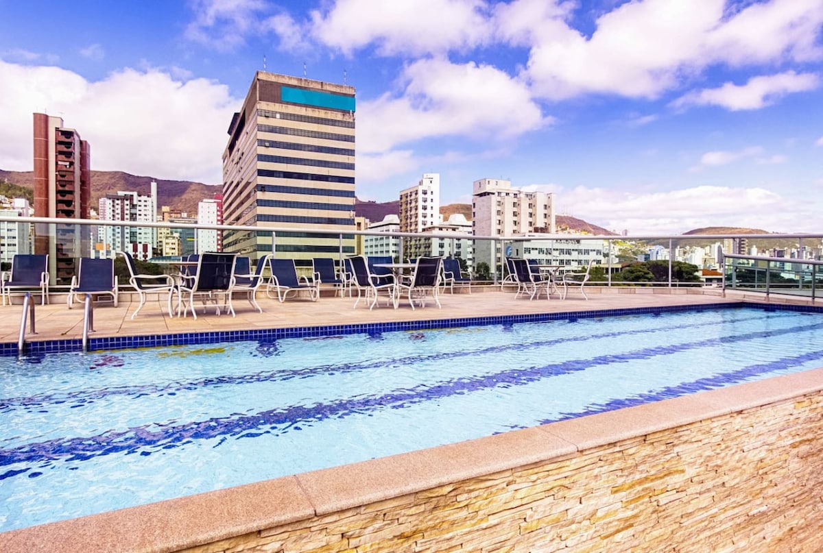An outdoor pool is visible, showcasing clear blue water and a stone border. Lounge chairs are arranged along the poolside. In the background, various modern buildings are seen against a vibrant sky, with mountains in the distance.