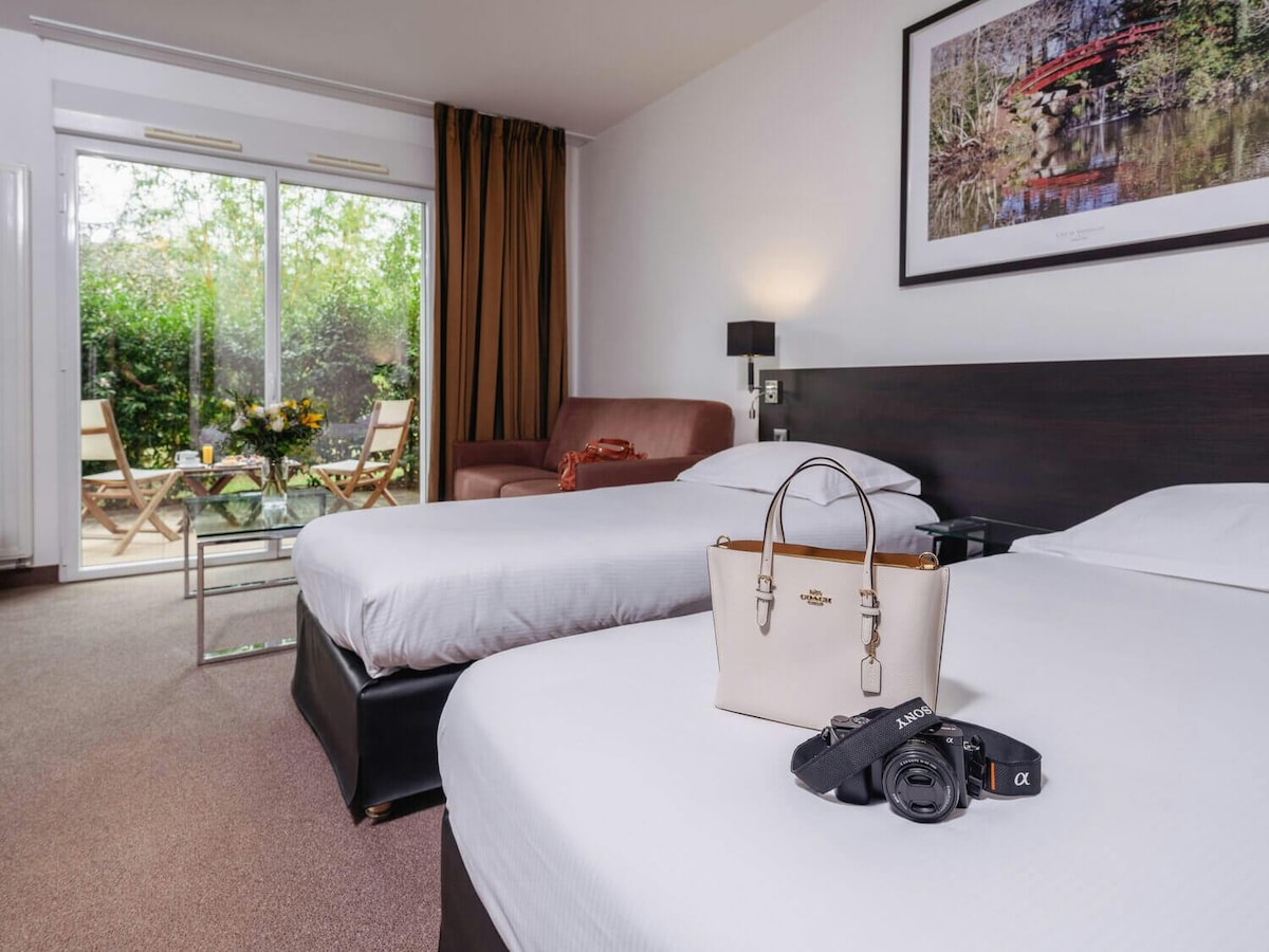 A well-lit studio features two single beds with crisp white linens, a convertible sofa, and a private balcony. A glass coffee table sits between the beds, and a camera and handbag are placed on one bed, showcasing the room's inviting atmosphere.