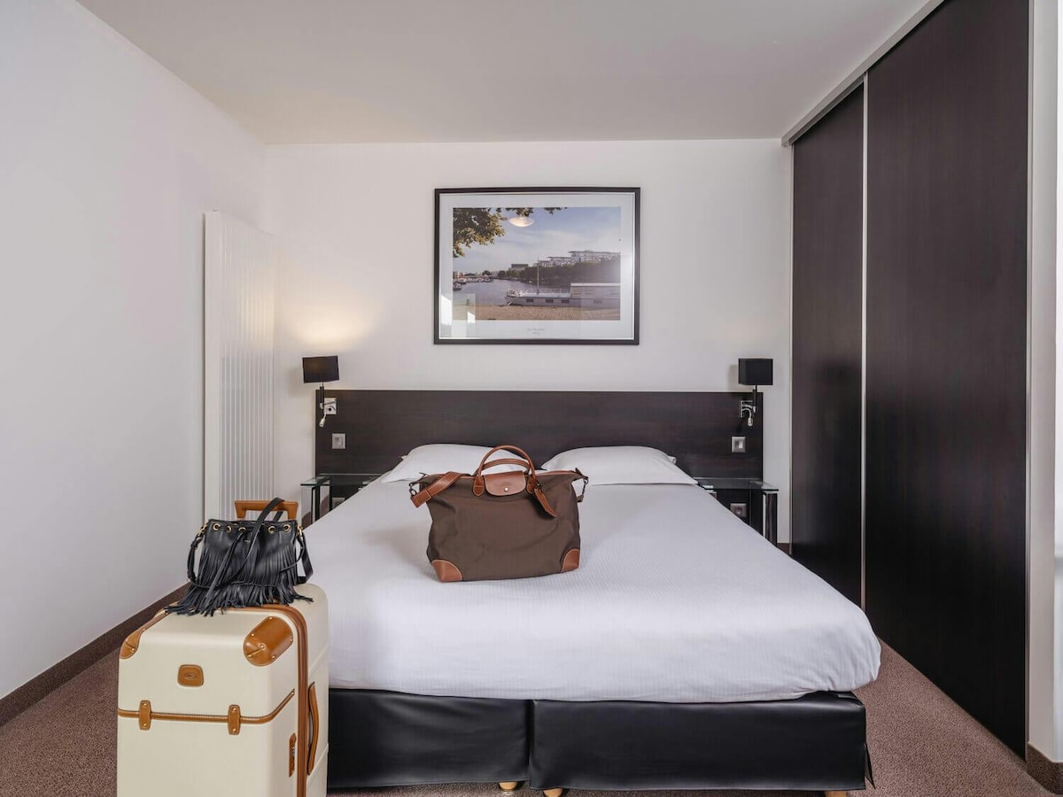 A spacious sleeping area features a large bed with crisp white linens. Luggage and a handbag are placed at the foot of the bed. On the wall hangs a framed black and white photograph, accompanied by modern bedside lamps. A sleek wardrobe can be seen to the right.