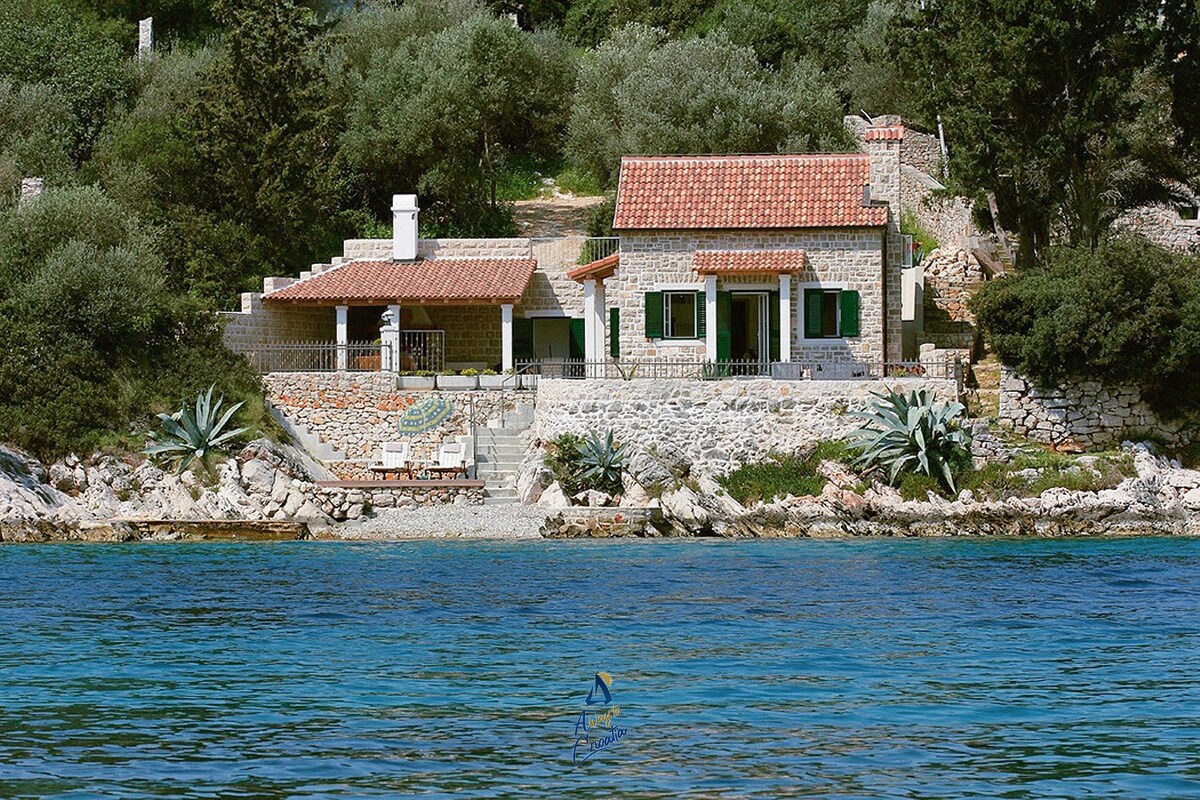 A charming stone house is situated along the waterfront, surrounded by lush greenery and olive trees. Large green-shuttered windows and a tiled roof complement the rustic architecture. A private patio offers views of the clear blue water, enhancing the coastal tranquility of the setting.