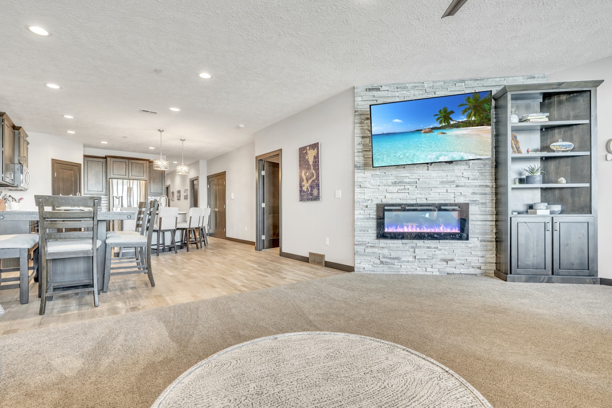 The living area is bright and open, featuring a modern electric fireplace beneath a wall-mounted television. A comfortable seating arrangement is highlighted by a large circular area rug. The space seamlessly connects to the kitchen and dining areas, creating an inviting environment for gatherings.