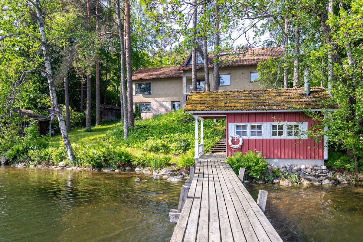 A charming lakeside scene features a wooden pier leading to a small boathouse with a red facade and a life preserver. The surrounding greenery includes lush trees and shrubs, complementing the serene lake in the foreground, with the house visible in the background.
