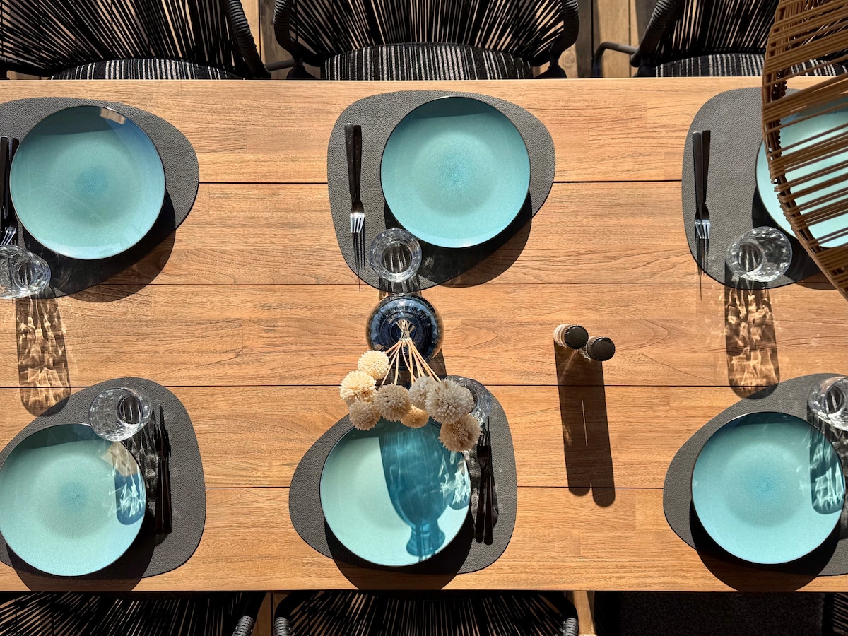 A large dining table is set for six, featuring round teal plates and cutlery. A decorative centerpiece with light-colored elements is placed at the center, enhancing the visual appeal. The table is surrounded by modern black chairs, providing a comfortable dining experience.