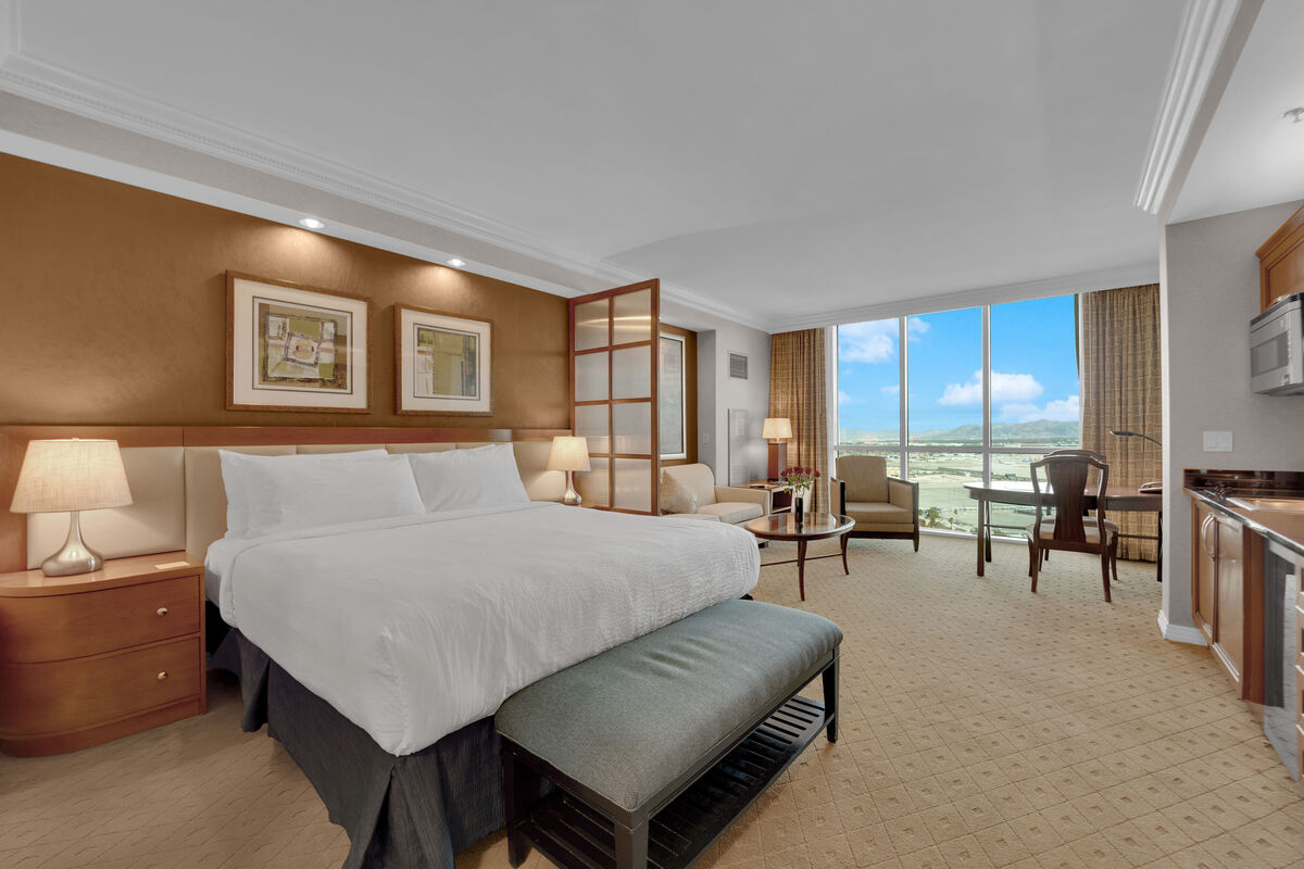 A spacious studio is filled with natural light from the large window, showcasing skyline views. A plush king bed dressed in white linens is positioned centrally, flanked by two bedside tables with lamps. A seating area with a sofa and coffee table offers relaxation, while a mini kitchen is included.