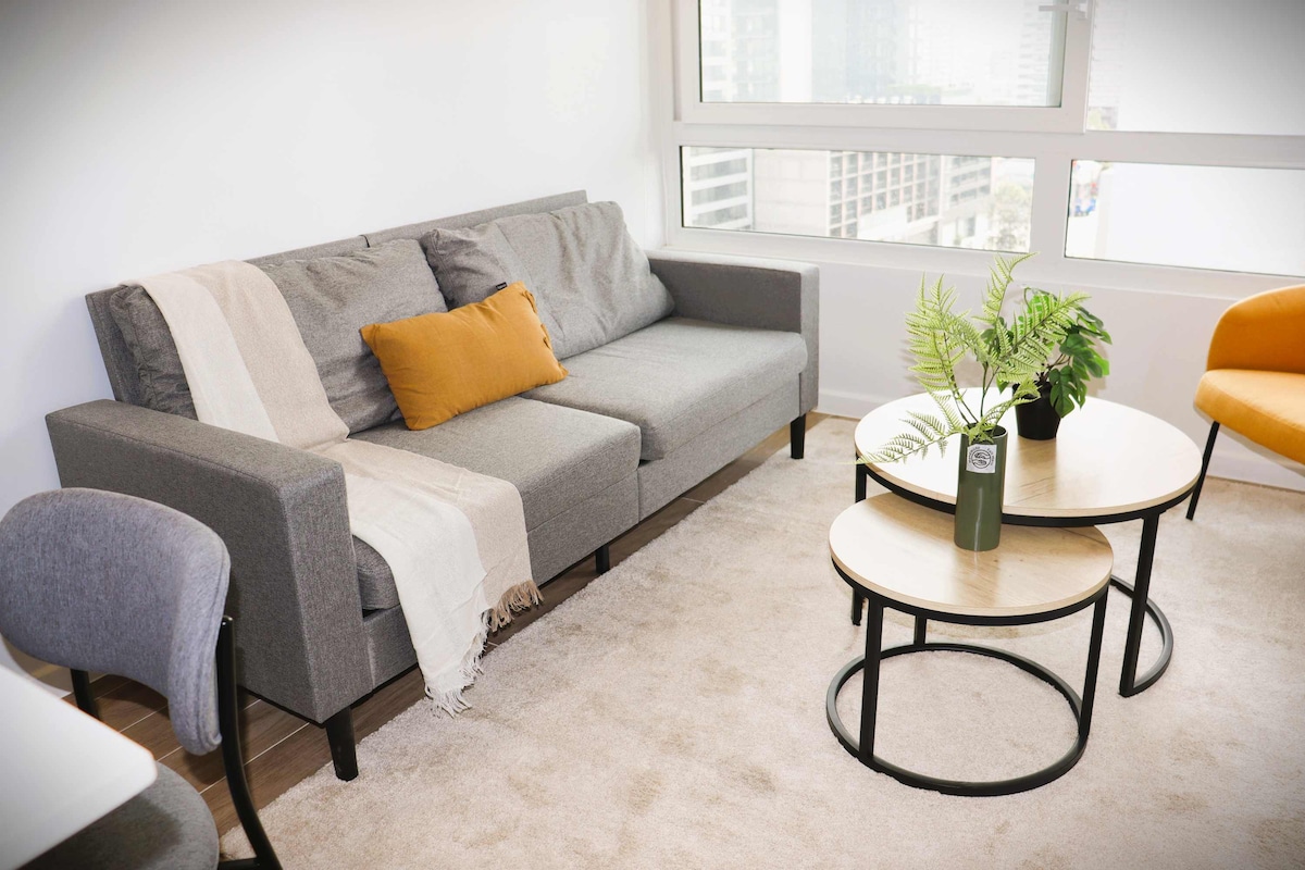 A modern living area features a gray sofa adorned with a warm-colored throw pillow. Two round coffee tables sit on a light carpet, accompanied by a potted plant. Natural light floods the space through large windows, enhancing the bright and airy ambiance.