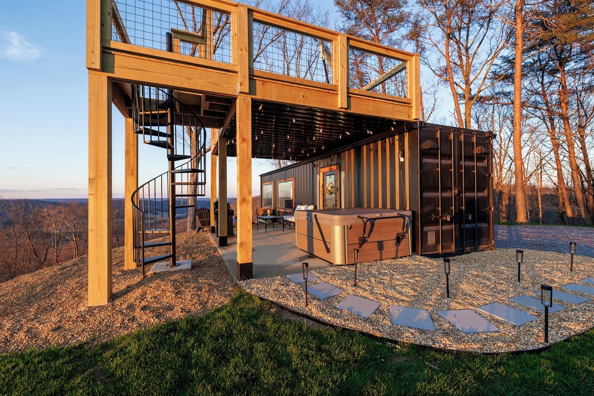 A unique retreat features a shipping container structure alongside a wooden deck elevated above ground. A spiral staircase leads to the deck, while a private hot tub is situated on the patio. Soft landscaping and outdoor lighting enhance the serene surroundings.