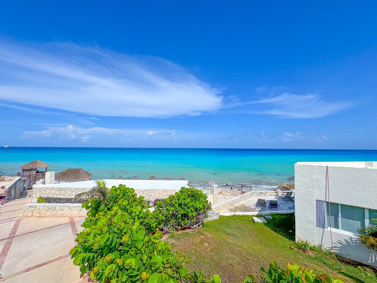 A panoramic view captures the clear turquoise sea meeting the horizon under a bright blue sky. The shoreline features beach loungers nestled among local greenery. The image showcases a sandy area with several palapas, suggesting a tranquil beach atmosphere.