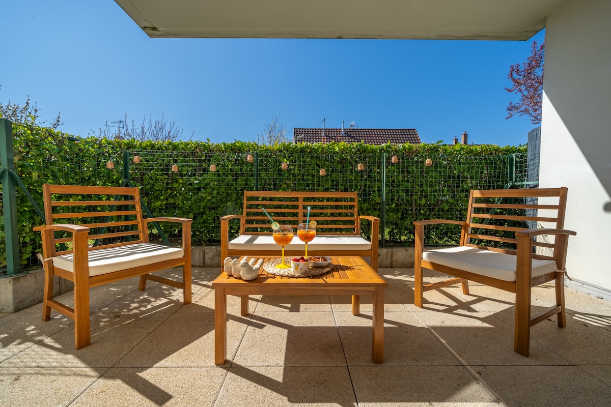 A private terrace is showcased with four wooden chairs and a table arranged for relaxation. Two glasses filled with orange beverages sit on the table, alongside a plate of snacks. Green foliage borders the space, providing a sense of greenery and privacy.