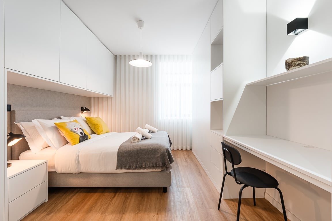 A modern bedroom features a cozy double bed dressed in white linens, complemented by accent pillows. A desk with a black chair is positioned next to the bed, along with built-in cabinetry. Soft natural light filters through sheer curtains, enhancing the serene atmosphere.