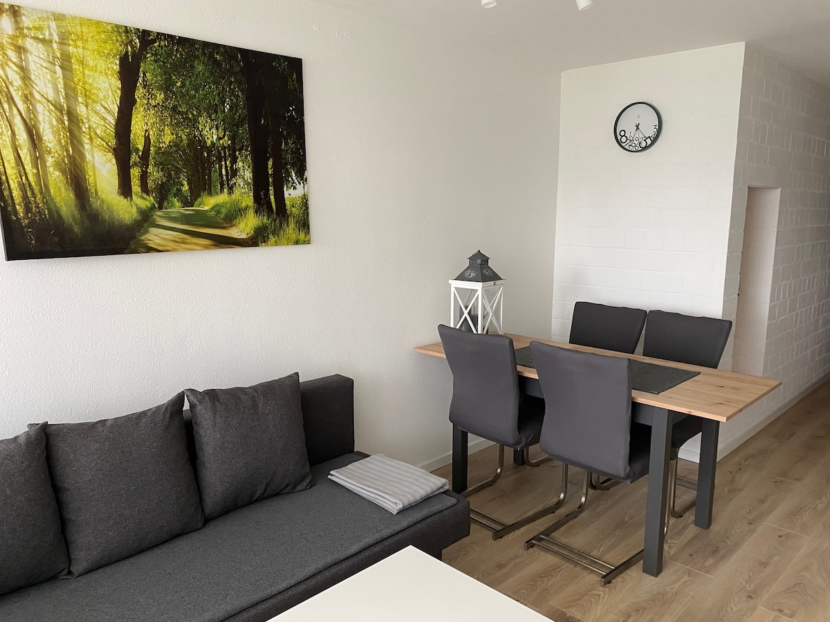 A bright living area features a gray sofa with a single cushion and a wooden dining table surrounded by four chairs. A large artwork depicting a sunlit path through trees adorns the wall, while a clock is mounted in the corner.