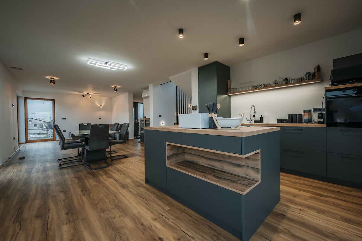 An open-concept kitchen and living area is highlighted by a central island featuring a wooden countertop. Modern appliances and minimalist cabinetry complement the spacious design, while ample natural light pours in through large windows. Comfortable seating is arranged for relaxation and socializing.