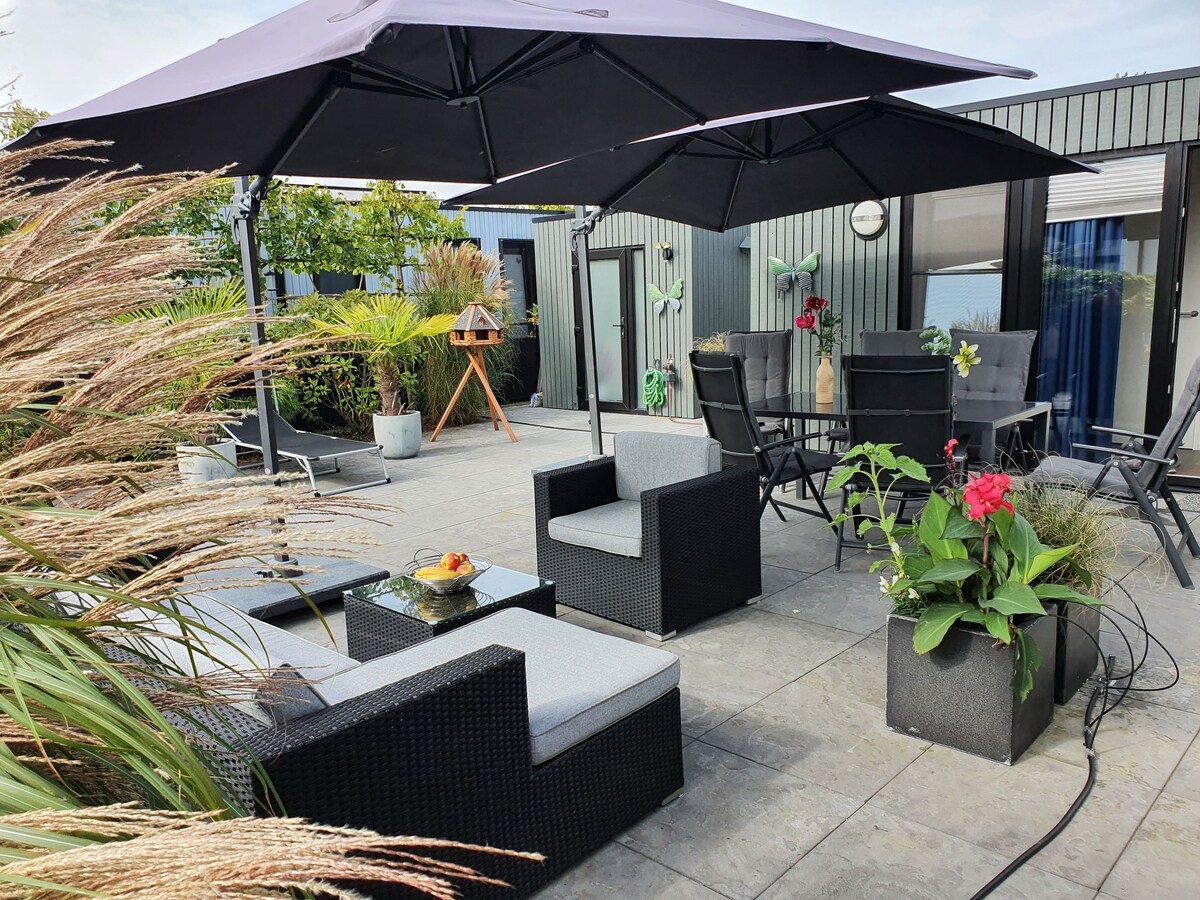 A spacious terrace is equipped with comfortable seating arranged around a table under large umbrellas. Decorative plants and flowers add vibrancy, while a barbecue grill is positioned nearby for outdoor cooking. The area offers a private setting for relaxation and social gatherings.