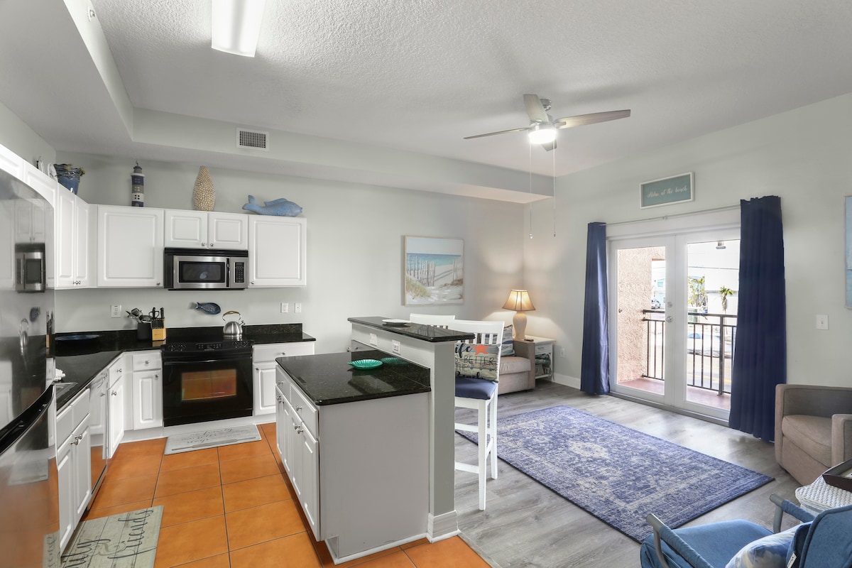 A spacious kitchen is visible, featuring contrasting black countertops and white cabinetry. A ceiling fan hangs above, while natural light enters through the sliding glass door leading to the private balcony. A cozy seating area with a blue rug is situated nearby.