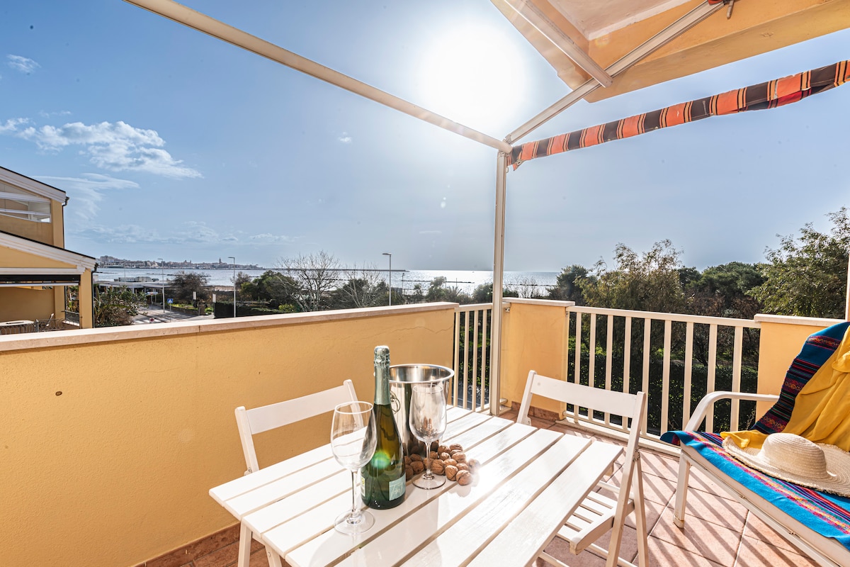 A sunlit balcony features a white dining table with two chairs, set with a bottle of wine and glasses. Scenic views of the sea and coastline can be seen in the background, while a vibrant blanket adds a splash of color to the setting.