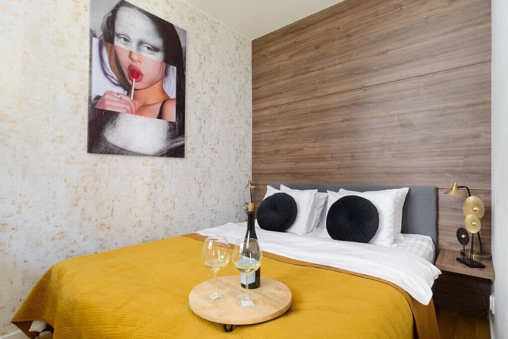 A comfortable bedroom is highlighted by a cozy bed dressed in soft linens and decorative pillows in a neutral palette. A wooden accent wall adds warmth, while a round tray with glasses and a bottle of sparkling water sits invitingly on the bed.