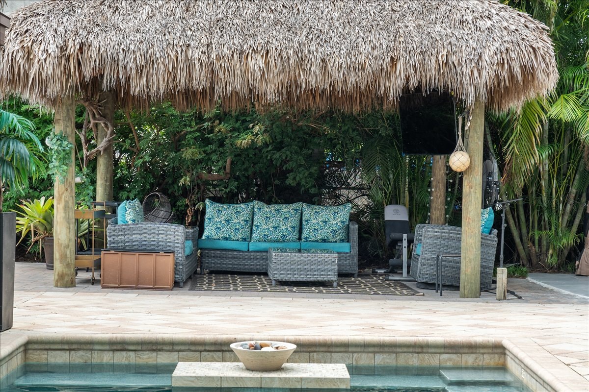 A shaded outdoor seating area is created under a thatched tiki hut, featuring a comfortable sectional sofa adorned with vibrant blue cushions. Lush greenery surrounds the space, providing a tropical backdrop, while a glimpse of the pool reflects the calming ambiance.
