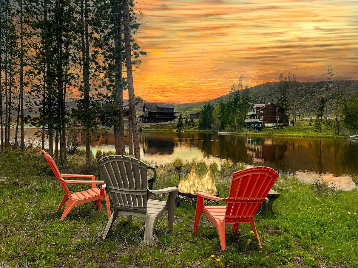 An outdoor seating area features three comfortable chairs arranged around a fire pit, with a serene lake reflecting a sunset filled with warm hues. Tall trees and lush vegetation surround the space, enhancing the tranquil atmosphere of the setting.