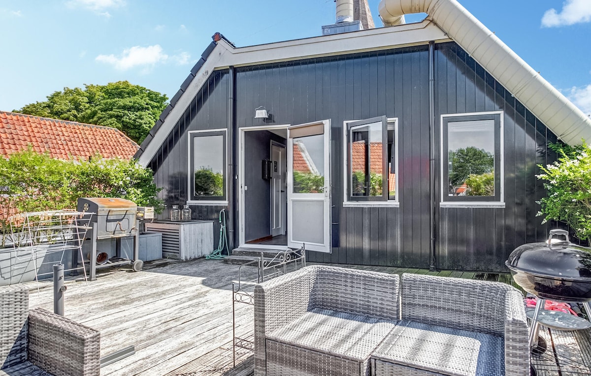 The spacious terrace features comfortable lounge furniture and a barbecue, surrounded by lush greenery. Two large windows provide ample natural light, and the entrance is marked by a welcoming doorway. The smooth wooden deck enhances the outdoor relaxation space.