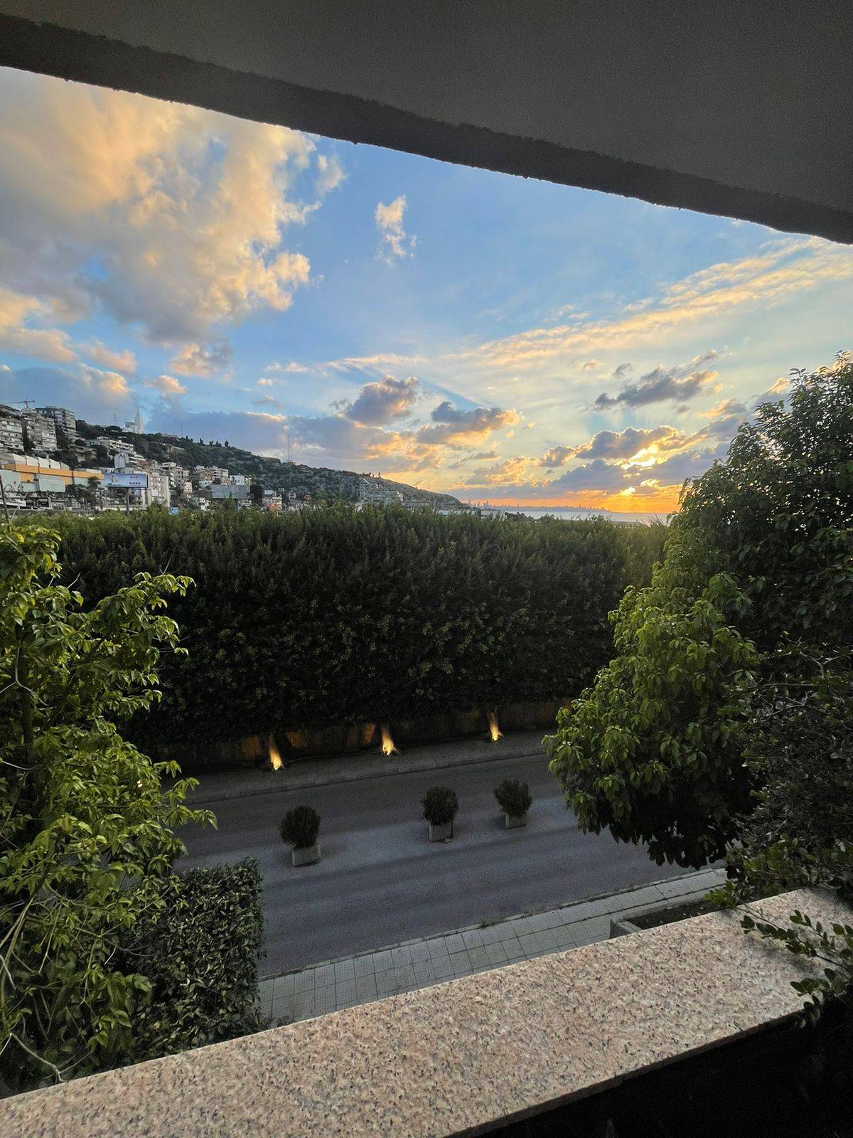 A balcony view showcases a sunset over a scenic landscape, with clouds in soft hues against the sky. Lush trees line the view, creating a natural frame. The street below is subtly illuminated, enhancing the tranquil atmosphere of the surrounding area.