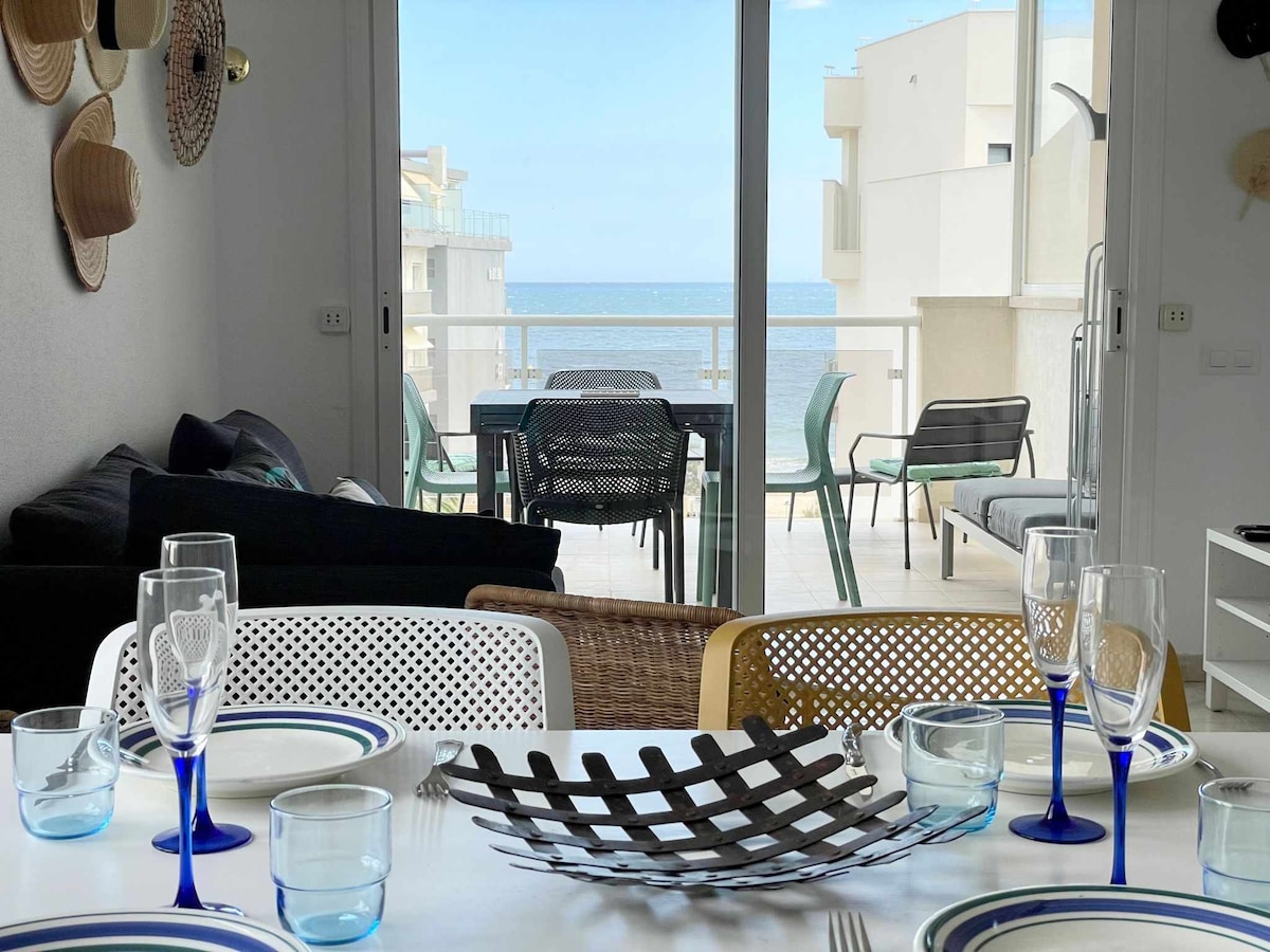A dining area is set for meals, featuring a white table with blue and patterned dishware. Glassware with blue stems sits beside a decorative black basket. A view of the outdoor terrace and the ocean is visible through large glass doors.