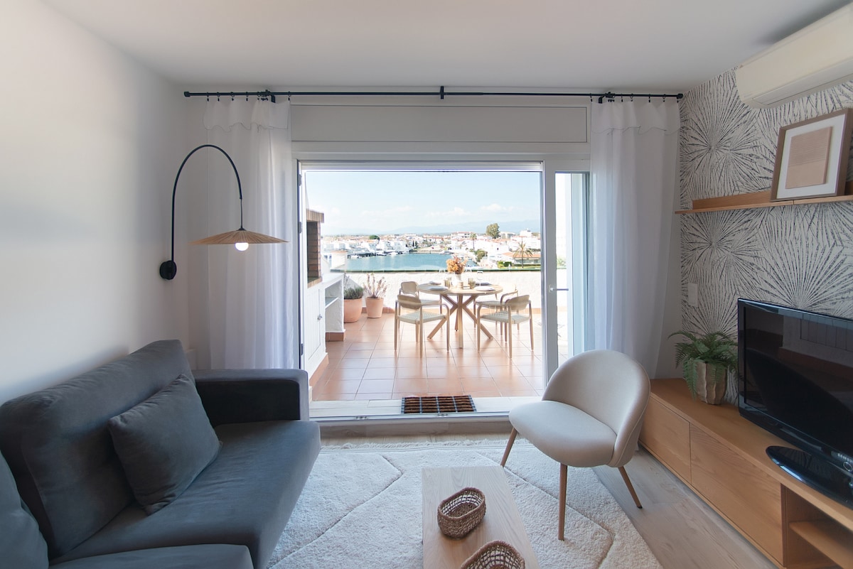 A living room area features a comfortable sofa and an armchair, complemented by a wooden coffee table. Large sliding doors provide access to a terrace with views of the canal. Soft curtains frame the doors, allowing natural light to fill the space.