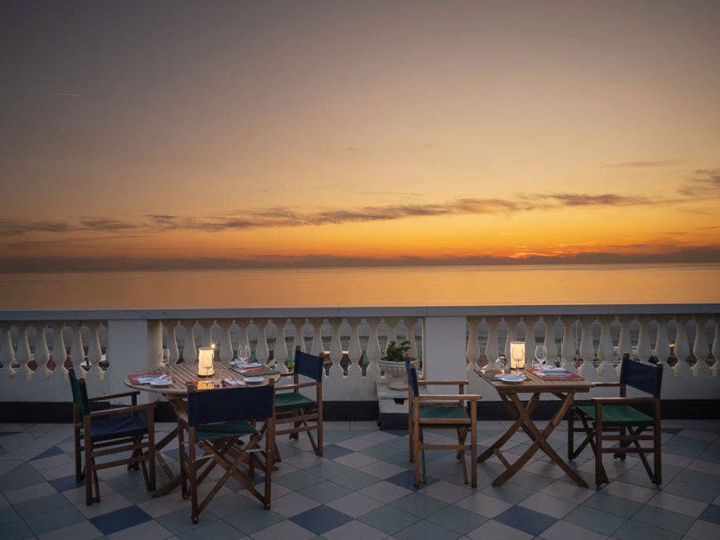 An open-air terrace features a set of wooden dining tables and chairs, arranged for casual dining. Soft lighting from lanterns enhances the tranquil ambiance as a colorful sunset paints the sky over the calm sea, creating a serene backdrop.